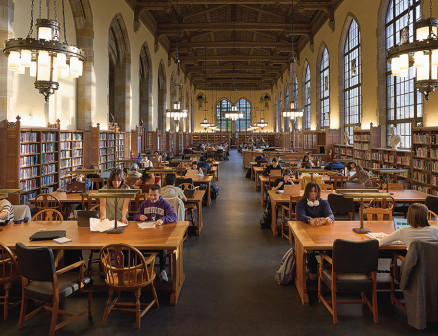 Students study at tables in a large light-filled room lined with bookshelves, with chandeliers above.
