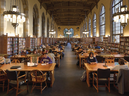 Students study at tables in a large light-filled room lined with bookshelves, with chandeliers above.