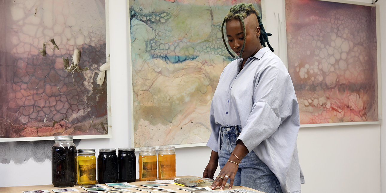 Artist Renee Royale stands at a table covered in Polaroids and jars of water; large works of art hang on the wall behind her.