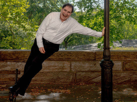 Richard Kind stands on the edge of a park bench with his arm outstretched as he leans over to his side, using his arm to brace himself against a streetlight on a stone walkway. He wears a white collared shirt and black slacks and is grinning at the camera. Trees are visible in the background.