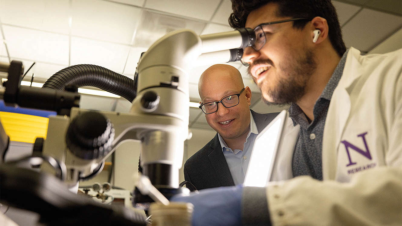 Juan Villacres Perez wears a lab coat and looks into a microscope while Jonathan Rivnay watches on in the background.