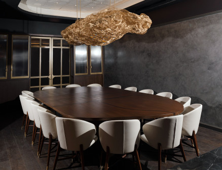 In a room with dark gray walls, 16 off-white chairs surround a dark wood oblong table and a sculptural chandelier hangs above.