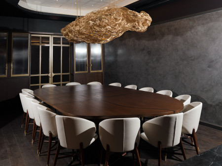 In a room with dark gray walls, 16 off-white chairs surround a dark wood oblong table and a sculptural chandelier hangs above.