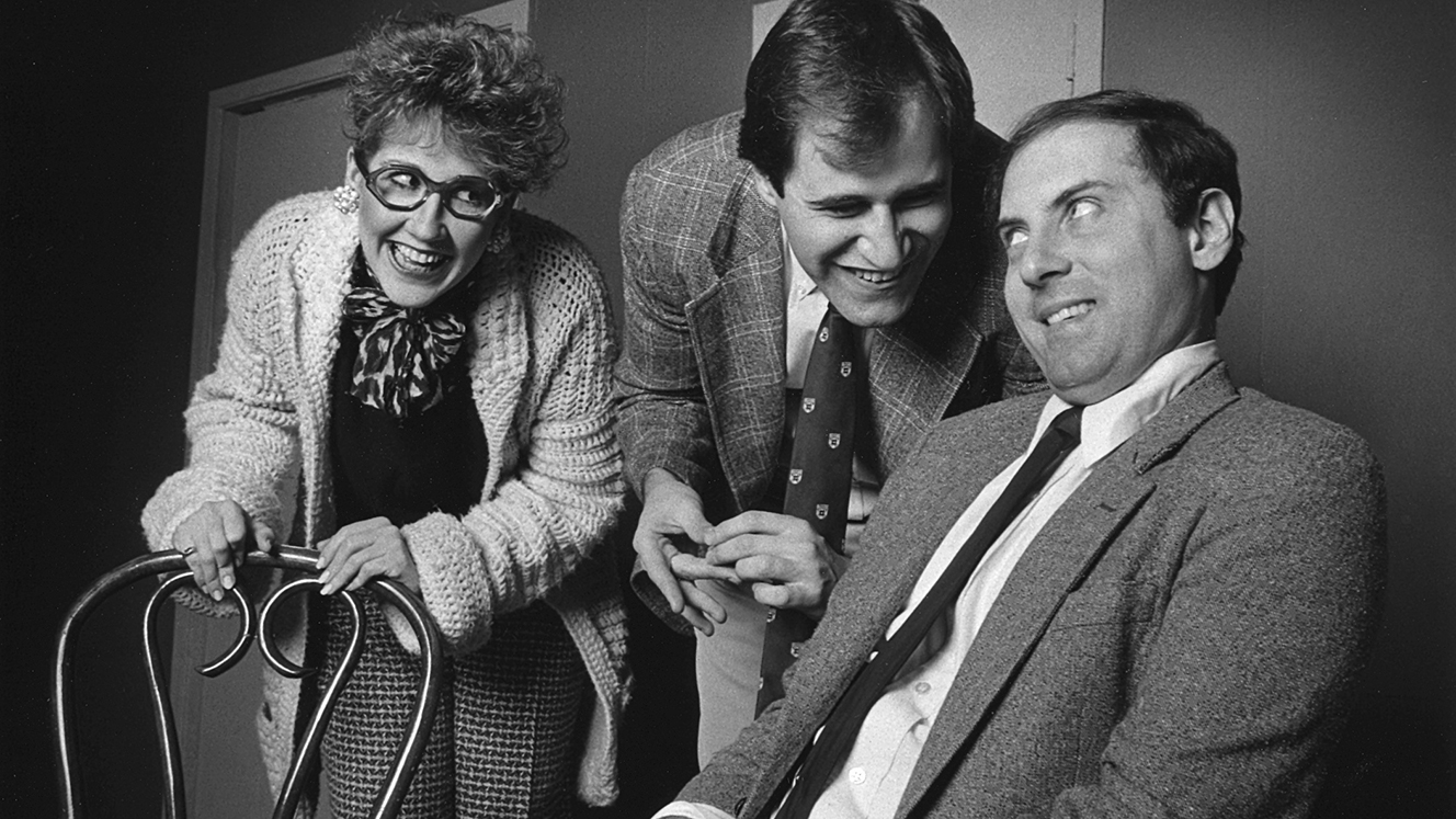 A scene from Second City’s 1986 main-stage revue Catch 27 featuring, in a row, a person holding onto the back of a chair and a person leaning over to grin at the person on the right, who is sitting.