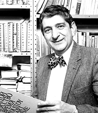 Irwin Weil wears a suit jacket with a bowtie while he reads a book and smiles at the camera.