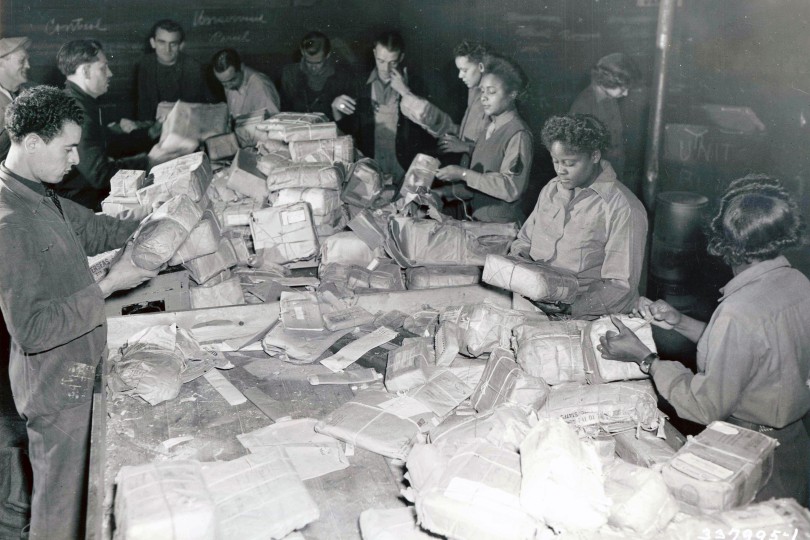 Black-and-white photo of women from the 6888th Central Postal Directory Battalion sorting through an enormous number of packages on a large table alongside French civilians.