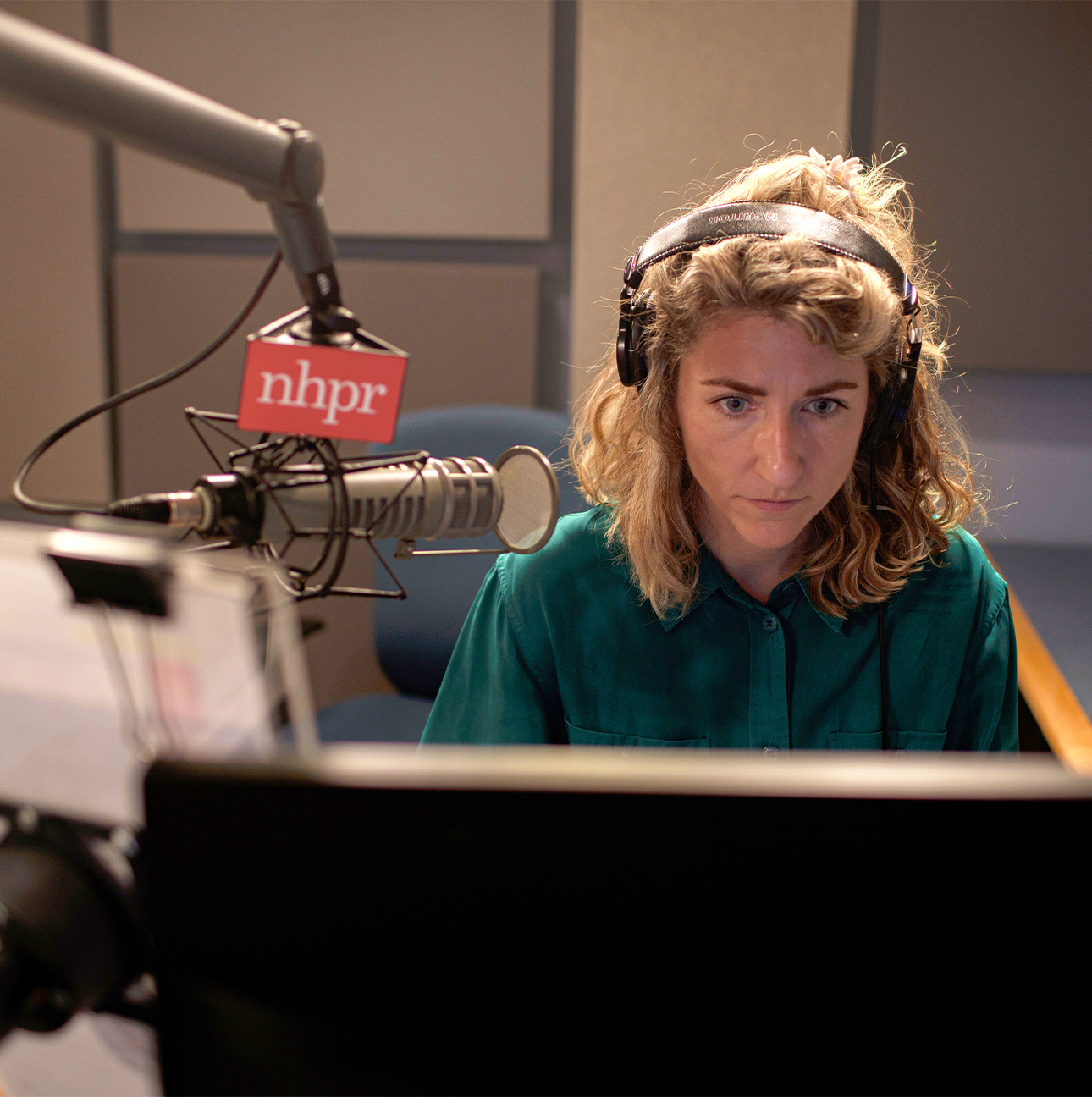 Lauren Chooljian wears headphones while sitting at a desk in a radio studio. There is a microphone at her right labeled “NHPR.”