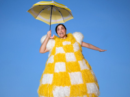 Liz Coin wears a huge, fuzzy, yellow-and-white checkered costume while holding a yellow and white umbrella against a blue background