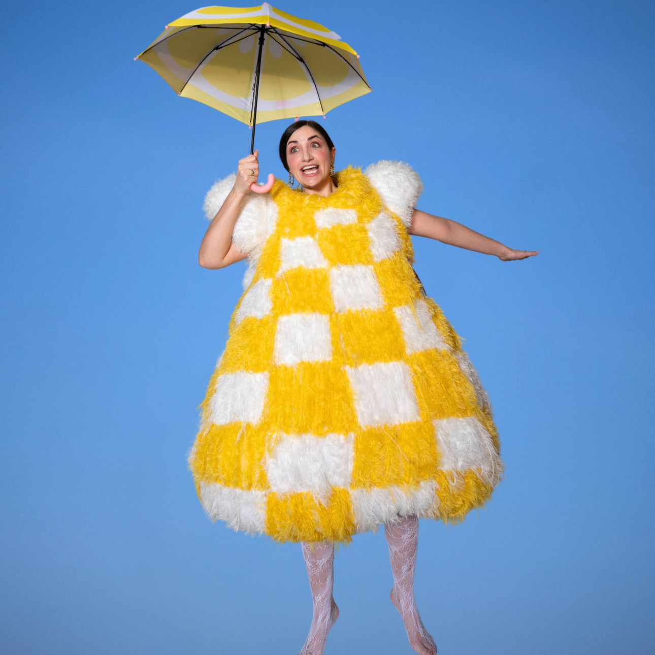 Liz Coin wears a huge, fuzzy, yellow-and-white checkered costume while holding a yellow and white umbrella against a blue background