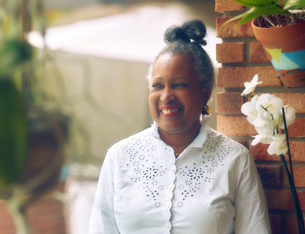 Monique Clesca leans up against a wall in Miami. She is smiling and wearing a white blouse. Her hair is in a bun.