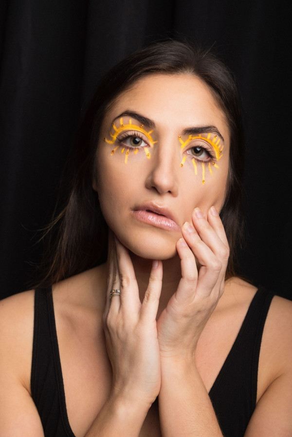 A headshot of Liz Coin wearing makeup around her eyes that look like rays of sun. She looks at the camera with a blank expression while her hands pull down on her face.