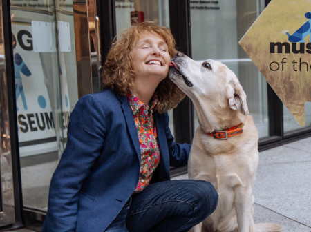 A woman poses with her yellow labrador retriever outside a building with a sign that reads, The AKC Museum of the Dog.