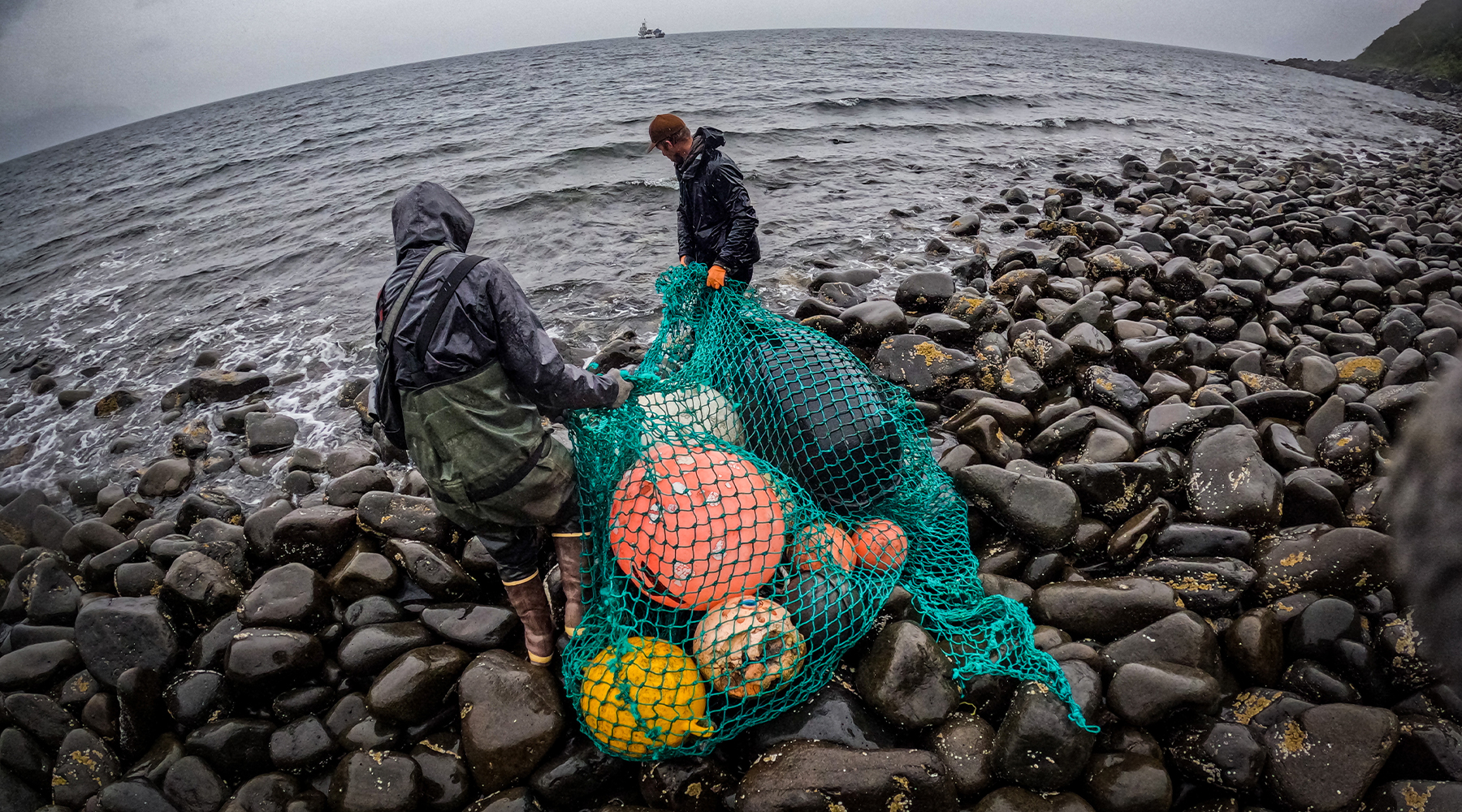 A crew of two people assemble marine debris for transport to the mother ship. They're pulling a bag of in this case derelict commercial fishing gear, including buoys and floats. They are standing on a rocky shoreline in the rain. There is a boat in the distance. 